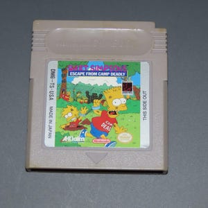 May include: A light gray Nintendo Game Boy cartridge featuring "Bart Simpson's Escape from Camp Deadly." The label shows Bart Simpson in a red shirt, blue shorts, and yellow skin. The cartridge is marked "DMG-TS-USA MADE IN JAPAN."