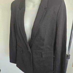 May include: A black blazer displayed on a mannequin. The blazer has a notched lapel, two front pockets, and a single-button closure. The sleeves are long, and the fabric appears to be a smooth, woven material. The blazer is a classic, versatile piece of clothing.