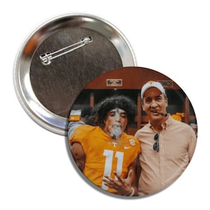 May include: A button featuring a photo of two men, one wearing a football jersey with the number 11 and the other wearing a plaid shirt. Both men are holding cigars.