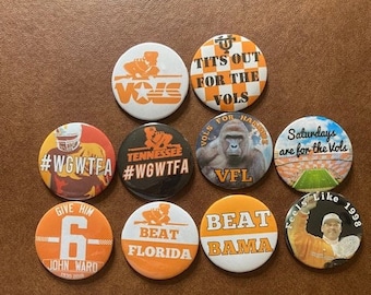 May include: Ten round buttons with various designs and slogans related to the University of Tennessee Volunteers football team. The buttons feature orange and white colors, with slogans such as "#WGWTFA", "VFL", "Beat Bama", and "Tits Out for the Vols".