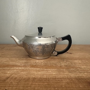 May include: A silver teapot with a black handle and lid knob. The teapot has an embossed design around the body. The teapot is on a wooden surface.