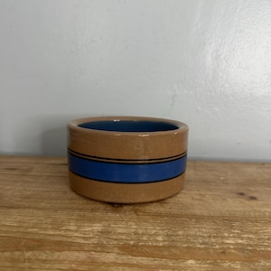 May include: A cylindrical ceramic bowl with a tan exterior and a blue interior. The exterior features a wide horizontal blue band flanked by two thin black stripes encircling the middle of the bowl, creating a simple and clean design.