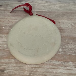 May include: A round, off-white ceramic ornament with a red ribbon for hanging. The ornament has the text "Pennsylvania Heart" and other text around the edge. The ornament is on a wooden surface.