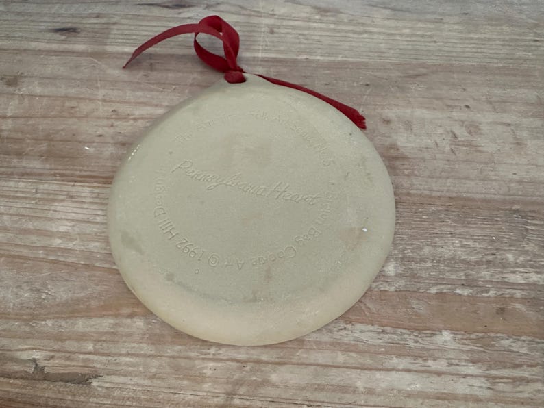 May include: A round, off-white ceramic ornament with a red ribbon for hanging. The ornament has the text "Pennsylvania Heart" and other text around the edge. The ornament is on a wooden surface.