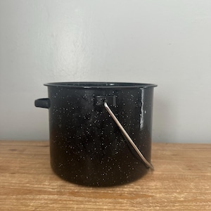 Op de afbeelding: Een zwarte emaille kampeerketel met witte spikkels. De ketel heeft aan elke kant een handvat en een metalen draadhandvat dat over de bovenkant boogt. De ketel staat op een houten oppervlak.