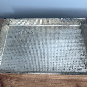 May include: Rectangular, vintage metal baking pan with a textured surface. The pan has slightly raised edges and shows signs of wear. The text "OVENEX" is visible on the pan. The pan is sitting on a wooden surface.