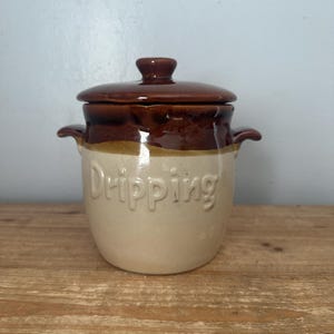 Könnte beinhalten: Ein Keramik-Tropfgefäß mit Deckel. Das Gefäß ist zweifarbig, mit einem cremefarbenen Boden und einem braunen Deckel. Das Wort "Dripping" ist auf der Vorderseite geprägt. Das Gefäß hat zwei kleine Griffe an den Seiten.