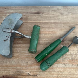 Op de afbeelding: Een verzameling vintage keukengereedschappen, waaronder een grote, gebogen metalen hakker met een groene handgreep en twee kleinere gereedschappen met groene handgrepen. De gereedschappen liggen op een houten oppervlak.