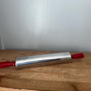 Può includere: Un mattarello d'argento con manici in plastica rossa. Il mattarello è su una superficie di legno.