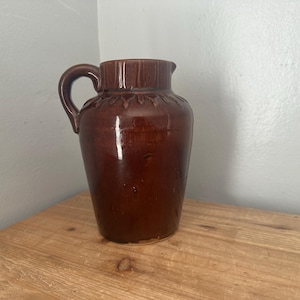 Op de afbeelding: Een bruine keramische kan met een handvat en een licht uitlopende tuit. De kan heeft een glanzende afwerking en een decoratieve band aan de bovenkant. Hij staat op een houten oppervlak.