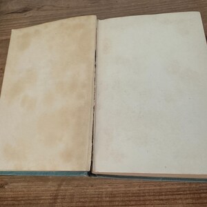 May include: An open book with aged, off-white pages. The left page has noticeable brown staining, while the right page is cleaner. The book's spine is visible, and the cover is a muted teal color. The book is lying on a wooden surface.