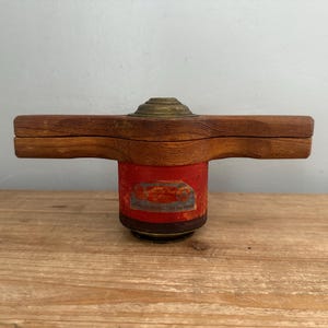 May include: A vintage wooden and brass object with a red cylindrical base. The wooden portion has a propeller-like shape. The red base has a faded label. The object is set on a wooden surface.