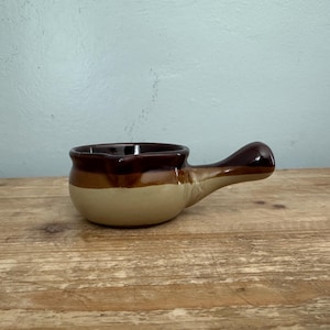 May include: A ceramic soup bowl with a single handle, featuring a two-tone glaze design. The upper portion of the bowl is a deep dark brown, while the lower section and the handle transition into a lighter, creamy beige color with a smooth finish.