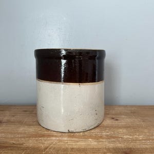 May include: A cylindrical stoneware crock featuring a two-tone design with a glossy dark brown glazed upper section and a matte off-white lower section. The vessel has a straight wall, a flat base, and a thick rim, showing signs of wear.