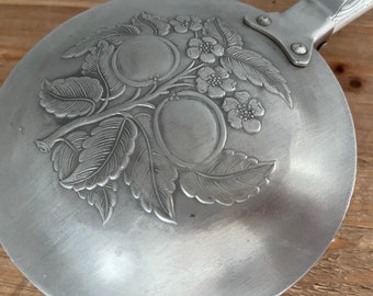 May include: A silver metal lid with an embossed design of leaves and fruit. The lid has a handle on the side.