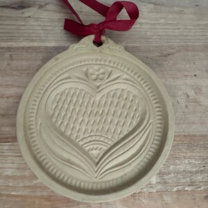 May include: A beige, round cookie mold with a heart design. The heart is textured with a woven pattern and surrounded by floral and leaf motifs. A burgundy ribbon is tied at the top for hanging. The mold is made of a light-colored material.