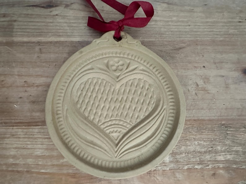 May include: A beige, round cookie mold with a heart design. The heart is textured with a woven pattern and surrounded by floral and leaf motifs. A burgundy ribbon is tied at the top for hanging. The mold is made of a light-colored material.