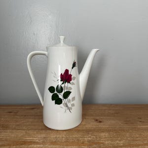 May include: White ceramic coffee pot with a lid and a handle. The pot is decorated with a red rose and green leaves. The pot is on a wooden surface.