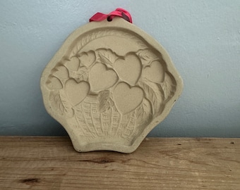 May include: Tan-colored ceramic cookie mold in the shape of a basket filled with hearts and leaves. A red ribbon is attached at the top for hanging. The mold is designed with detailed relief patterns, perfect for baking themed cookies.
