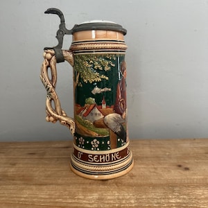 May include: A ceramic beer stein with a metal lid and a vine-shaped handle. The body features a relief scene of a person sitting in a landscape with houses and trees, with the text O' SCHÖNE ZU printed in white on a brown band at the base.