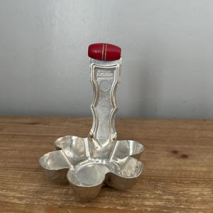 Op de afbeelding: Een zilverkleurige, bloemvormige serveerschaal met zes compartimenten. Het heeft een handvat met een rode, ovaalvormige bovenkant. Het handvat heeft de tekst "Silverplate" en "Patent". De schaal staat op een houten oppervlak.