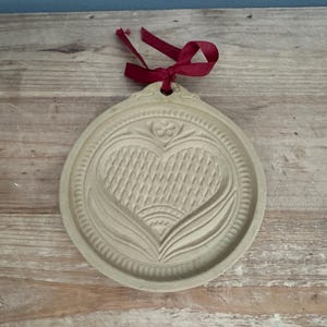 May include: A beige, round cookie mold with a heart and floral design. The heart has a woven texture, and the mold is trimmed with a decorative border. A red ribbon is attached at the top for hanging.