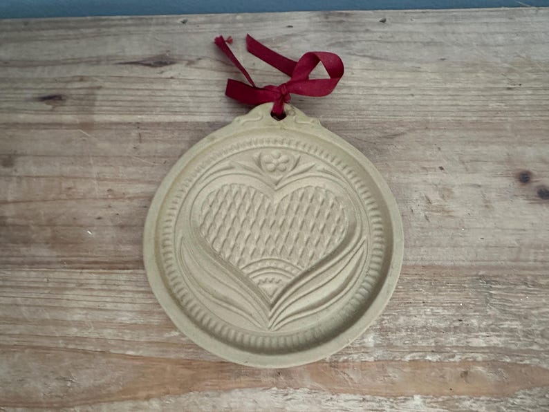 May include: A beige, round cookie mold with a heart and floral design. The heart has a woven texture, and the mold is trimmed with a decorative border. A red ribbon is attached at the top for hanging.