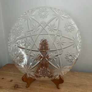 Può includere: Un piatto da portata rotondo in vetro trasparente con un motivo a stella, esposto su un supporto in legno. Il piatto presenta un design dettagliato di stelle e forme geometriche. Ideale per servire antipasti o dessert.