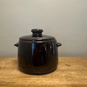 May include: A dark brown ceramic pot with a lid. The pot has two small handles on the sides and a rounded lid with a small knob. The pot is sitting on a wooden surface.