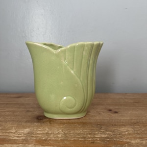 May include: A light green ceramic vase with a tulip-like shape. The vase has a fluted design on the sides and a decorative swirl. The vase is sitting on a wooden surface.