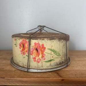 May include: A vintage round metal cake carrier with a cream-colored finish and a hand-painted floral design featuring pink and orange flowers with green leaves. The container shows signs of rust and includes a wire handle and a clear knob.