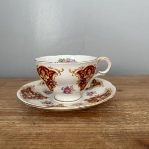May include: A vintage teacup and saucer set. The white porcelain cup and saucer feature a burgundy and gold floral design. The cup has a delicate handle and sits on a small pedestal. The saucer has a matching pattern around the rim.