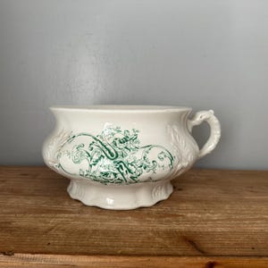 Op de afbeelding: Crèmekleurige keramische po met een groen bloemmotief. De pot heeft een handvat en een decoratieve voet. Het bloemmotief herhaalt zich rond de body. Het staat op een houten oppervlak.