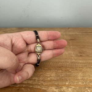 May include: A vintage-style wristwatch featuring a small, rectangular gold-tone case with a dark face and gold-colored numerals. The watch is attached to a thin, black cord strap with gold-tone metal connectors at the lugs, creating a delicate look.