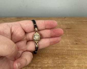 May include: A vintage-style wristwatch featuring a small, rectangular gold-tone case with a dark face and gold-colored numerals. The watch is attached to a thin, black cord strap with gold-tone metal connectors at the lugs, creating a delicate look.