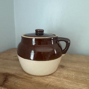 Puede incluir: Un tarro de cerámica de dos tonos con tapa. La parte superior es de color marrón oscuro brillante, mientras que la mitad inferior es de color crema. Tiene un asa y un pequeño pomo en la tapa. El tarro está sobre una superficie de madera.