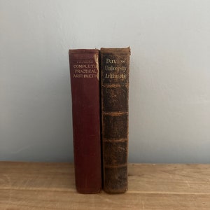 Może przedstawiać: Dwie stare książki w twardej oprawie ze zużytymi grzbietami. Książka po lewej jest czerwona i ma napis "Heath's Complete Practical Arithmetic". Książka po prawej jest brązowa i ma napis "Davies' University Arithmetic".