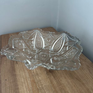 May include: Clear, decorative glass serving bowl with a scalloped edge. The bowl features a detailed, geometric pattern with starburst and leaf-like designs. The bowl is rectangular with rounded corners and sits on a wooden surface.