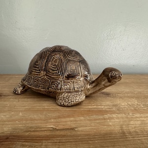 Könnte beinhalten: Eine braune Keramik-Schildkrötenfigur mit einem detaillierten Panzermuster. Die Schildkröte sitzt auf einer Holzoberfläche.