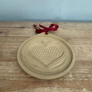 May include: A round, beige cookie mold with a heart design and a red ribbon. The heart is textured with a woven pattern and surrounded by a floral design. The mold is made of a light-colored material and sits on a wooden surface.