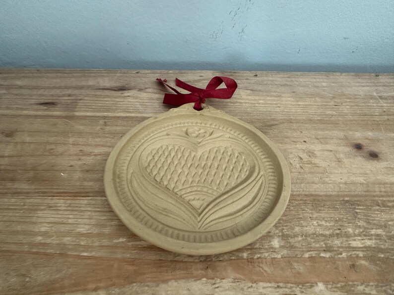 May include: A round, beige cookie mold with a heart design and a red ribbon. The heart is textured with a woven pattern and surrounded by a floral design. The mold is made of a light-colored material and sits on a wooden surface.
