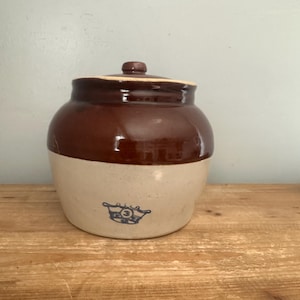 May include: A two-toned stoneware crock with a dark brown glazed upper section and a light beige lower section. The jar features a matching lid with a small knob and a blue stamped crown logo containing the number 3 on the front lower body.