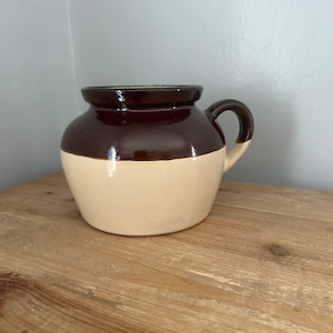 Puede incluir: Una jarra de cerámica esmaltada marrón y crema con asa. La jarra tiene un cuerpo redondeado y una boca ancha.