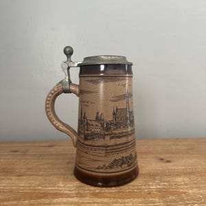 May include: A vintage ceramic beer stein with a hinged metal lid. The stein features a detailed sepia-toned illustration of a European cityscape. The handle and base are a darker brown, with a lighter tan body.