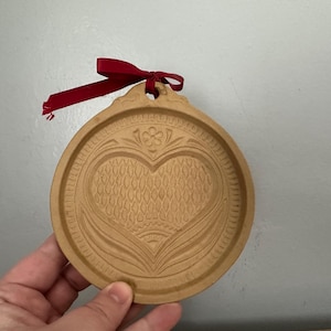 May include: A round, wooden ornament with a heart design carved into the surface. The ornament has a decorative border and a small hole at the top with a red ribbon tied through it. The ornament is light brown in color.