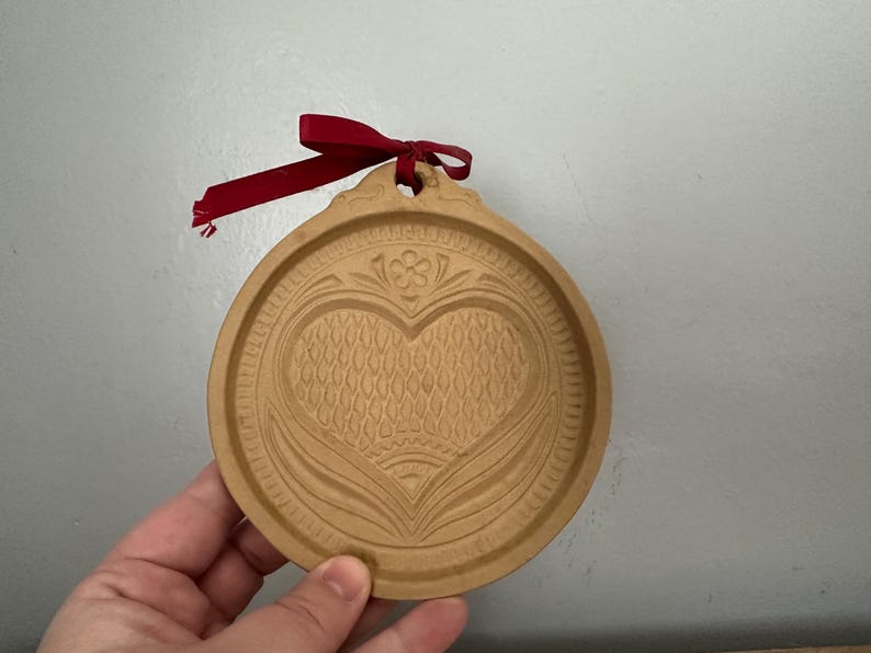 May include: A round, wooden ornament with a heart design carved into the surface. The ornament has a decorative border and a small hole at the top with a red ribbon tied through it. The ornament is light brown in color.