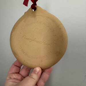 May include: A tan, ceramic Pennsylvania Heart ornament with a hole for hanging and a red ribbon. The back of the ornament features embossed text: "The American Folk Art Series No. 5" and "Brown Bag Cookie Co."