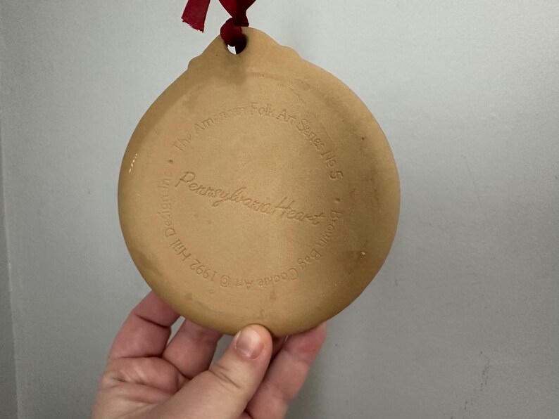 May include: A tan, ceramic Pennsylvania Heart ornament with a hole for hanging and a red ribbon. The back of the ornament features embossed text: "The American Folk Art Series No. 5" and "Brown Bag Cookie Co."
