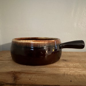 May include: A brown and white glazed ceramic serving dish with a handle. The dish has a rounded shape and a slightly flared rim.