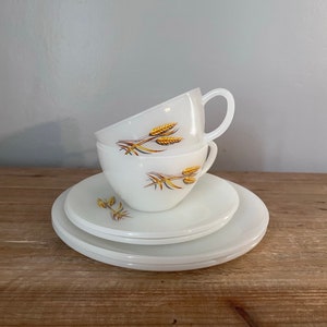Peut inclure: Deux tasses à thé en céramique blanche avec un motif de blé doré empilées l'une sur l'autre sur une soucoupe blanche. La soucoupe est posée sur une surface en bois.